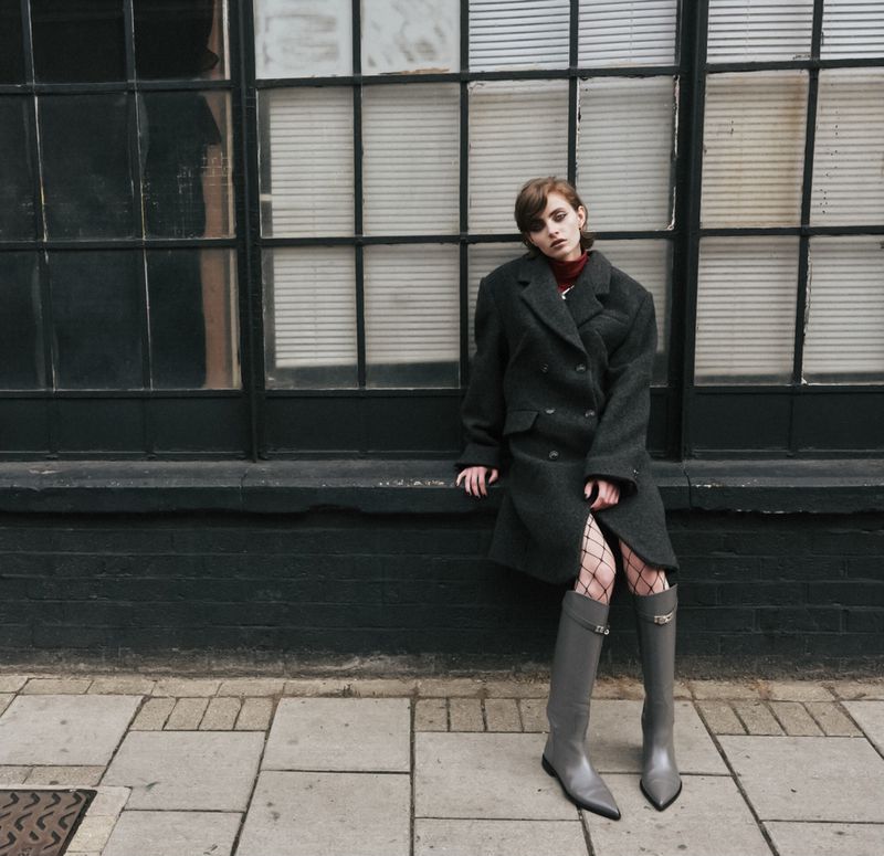 Woman in oversized coat and gray boots poses against a large black window. The sidewalk and brick wall add depth to the urban scene.