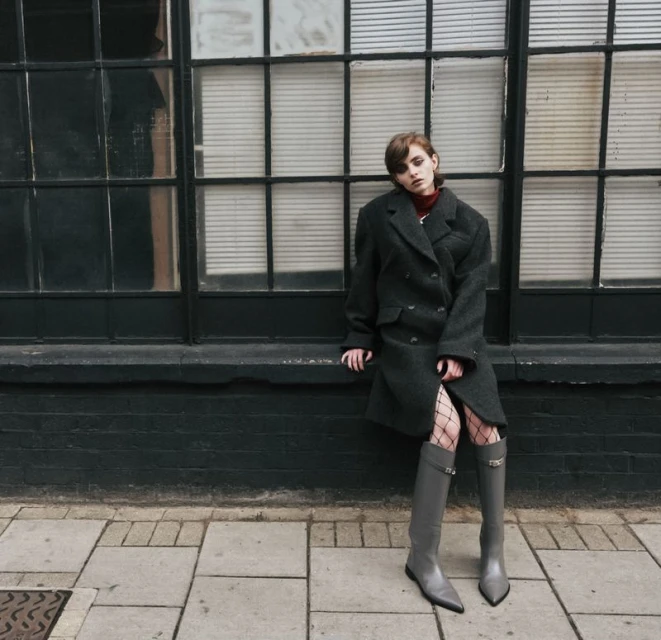 Woman in oversized coat and gray boots poses against a large black window. The sidewalk and brick wall add depth to the urban scene.