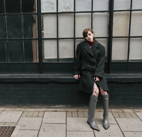 Woman in oversized coat and gray boots poses against a large black window. The sidewalk and brick wall add depth to the urban scene.