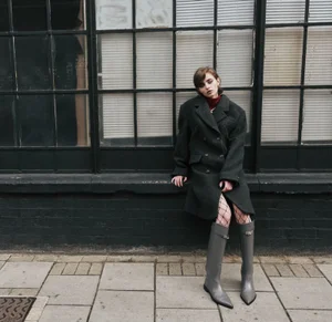 Model leaning against a dark framed industrial window wearing an oversized grey coat and knee high boots