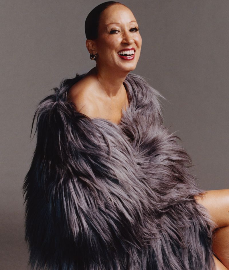 Attractive mature woman with short hair smiles broadly while posing indoors, wrapped in a gray faux fur coat against a gray background.
