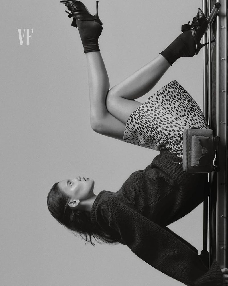 Upside-down view of a woman in a sweater and spotted skirt, wearing high-heeled shoes, posing against a metal frame in a studio.
