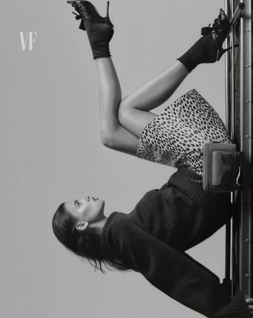 Upside-down view of a woman in a sweater and spotted skirt, wearing high-heeled shoes, posing against a metal frame in a studio.
