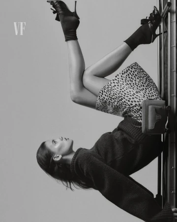 Upside-down view of a woman in a sweater and spotted skirt, wearing high-heeled shoes, posing against a metal frame in a studio.