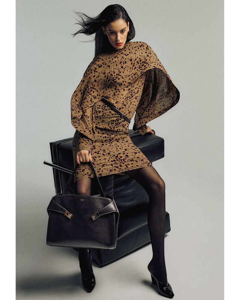 A woman sits on a black leather chair with a matching bag. She wears a brown animal-print dress, black tights, and high heels.