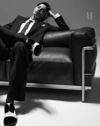 Model wearing a black suit and tie reclining on a leather sofa against a neutral background