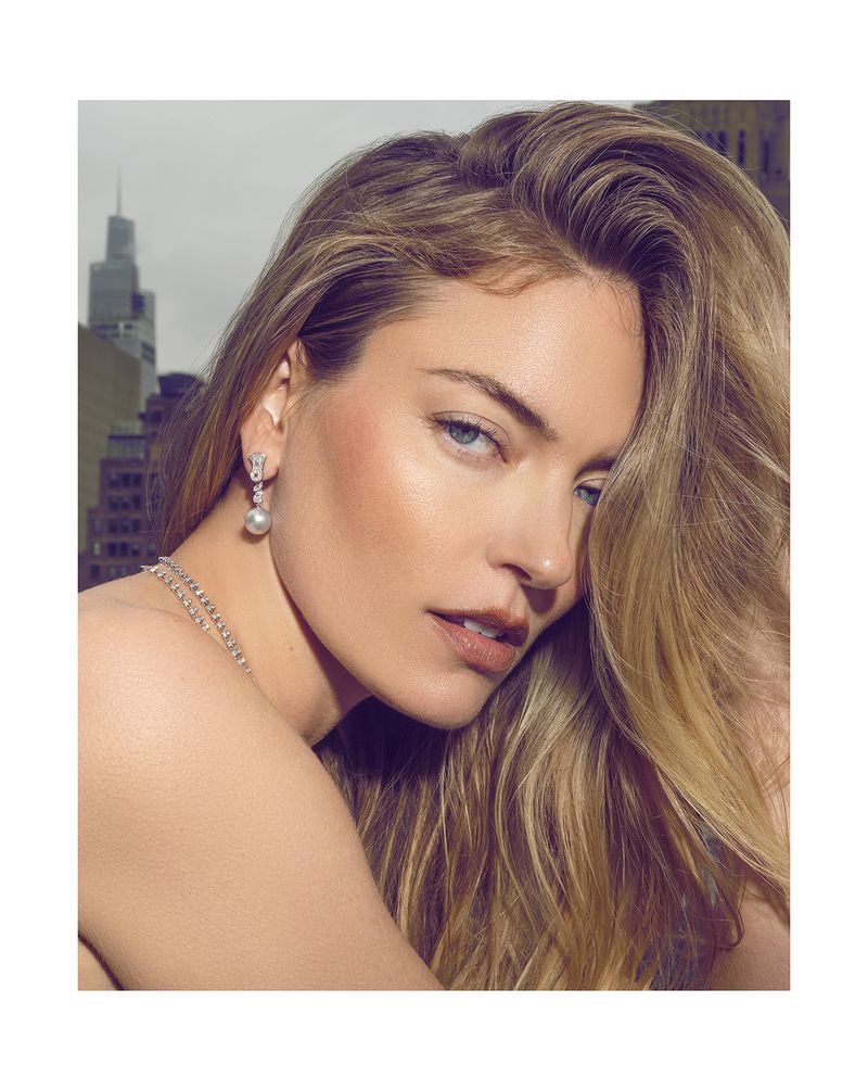 Close-up of a blonde woman with blue eyes wearing pearl drop earrings and a necklace, with an urban skyline visible in the background.