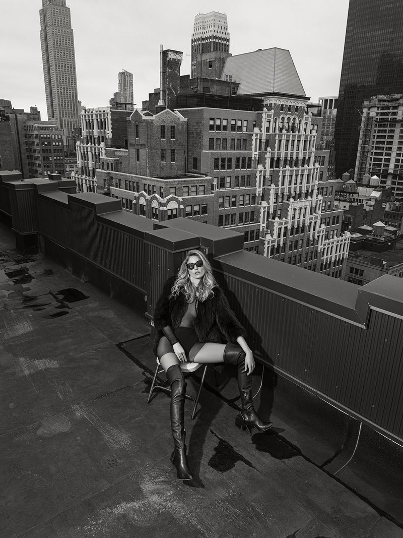 Fashion editorial shot of a woman in sunglasses, coat, and boots sitting on a chair on a rooftop with a cityscape in the background.
