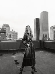 Model Martha Hunt wearing a long dark coat on a rooftop with city skyline backdrop