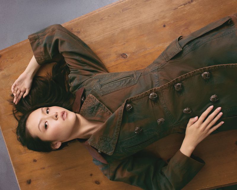 An Asian woman with long black hair is laying on a light brown wooden surface wearing a dark green buttoned coat and looking at the camera.