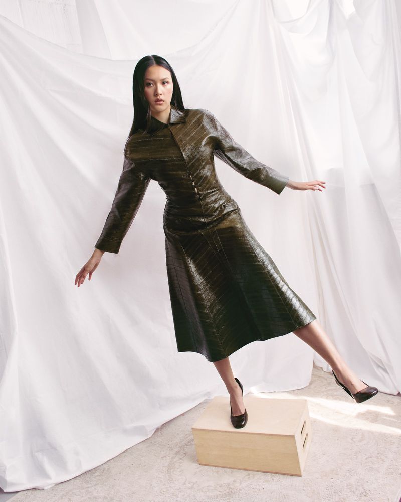 A stylish Asian woman stands on a wooden box in a light green reptile-patterned leather dress, with a flowing white cloth background.