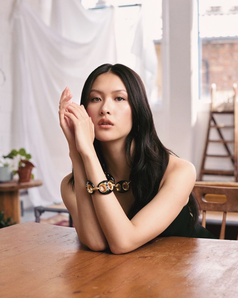 Elegant Asian woman in a black top poses at a wooden table, wearing a gold-and-black link necklace, with a soft, bright backdrop.