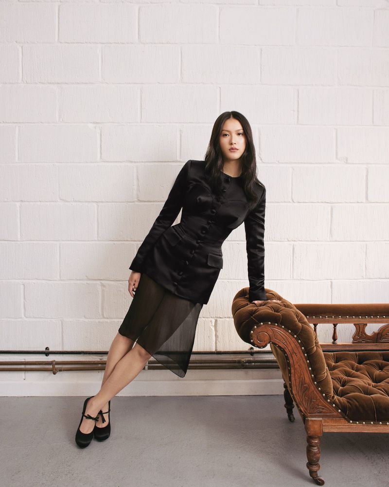 Elegant Asian woman in a black blazer dress leans on a vintage chaise lounge in front of a white brick wall for a fashion portrait.