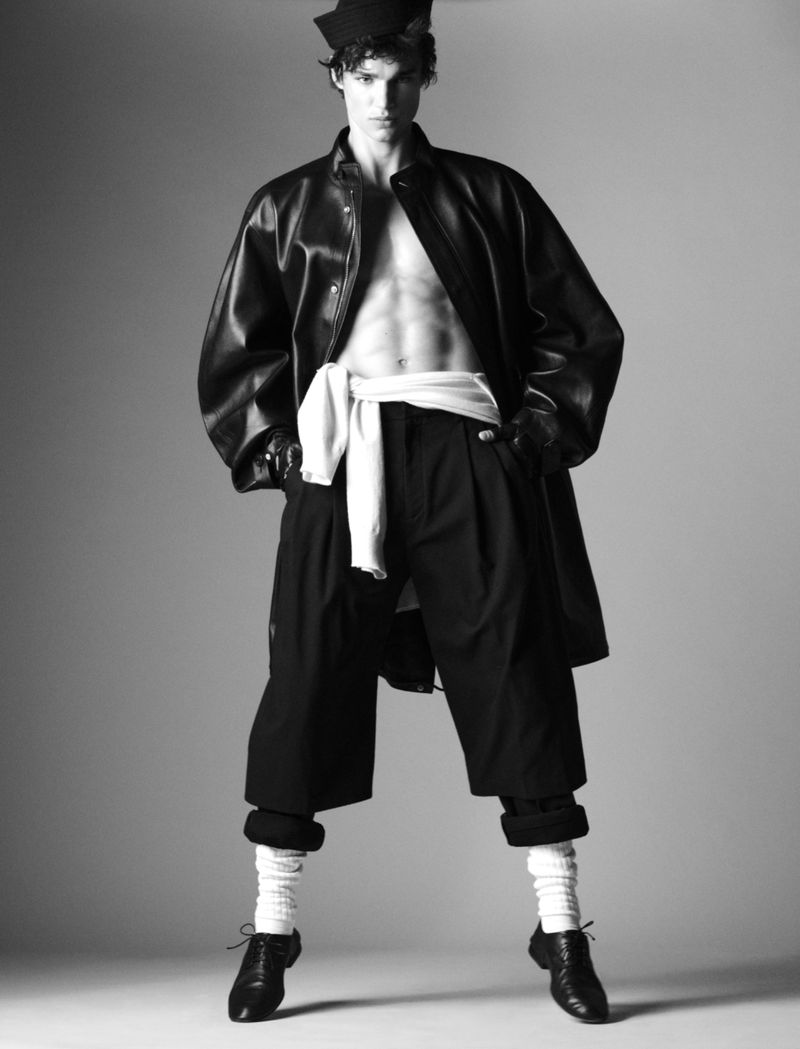 Black-and-white studio portrait of a male model in unbuttoned leather coat, abs visible, culottes, long socks, and black derby shoes.