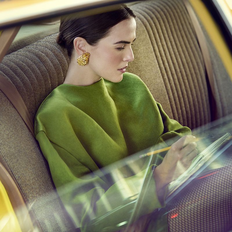 Elegant woman with jewelry inside a classic car sketches in a notebook. She wears a green top and poses next to the patterned interior.