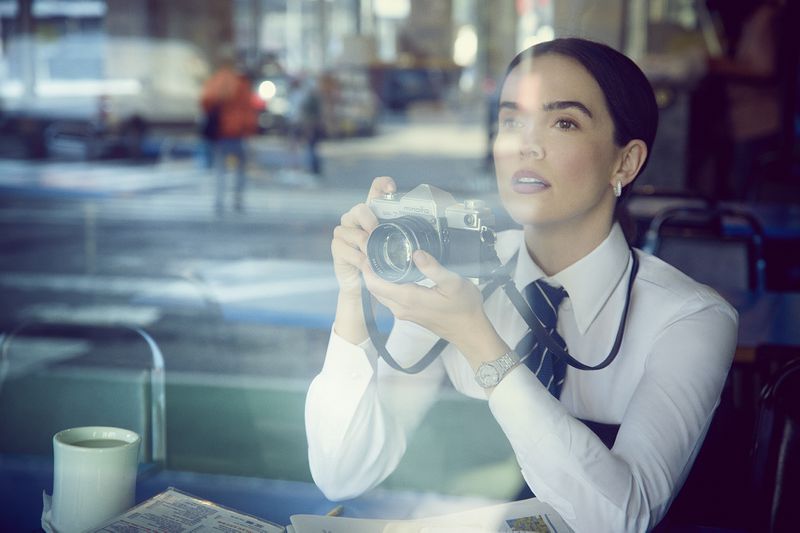 Woman in a white shirt and tie holding a vintage camera, looking up and out through a window in a city cafe setting.