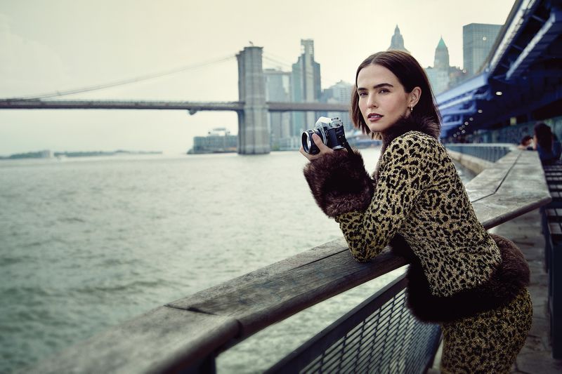 Woman in animal-print jacket holds camera, standing on a pier with the Brooklyn Bridge and New York City skyline in the background.