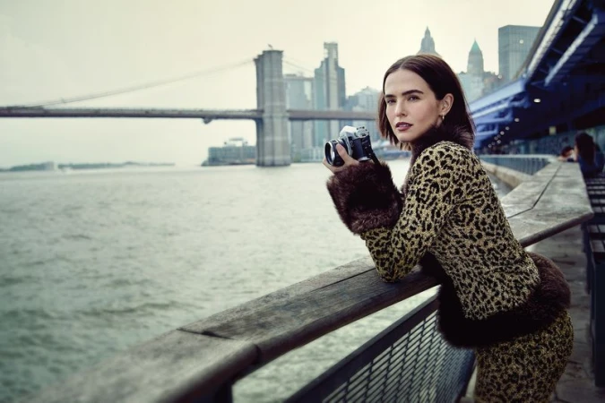 Woman in animal-print jacket holds camera, standing on a pier with the Brooklyn Bridge and New York City skyline in the background.