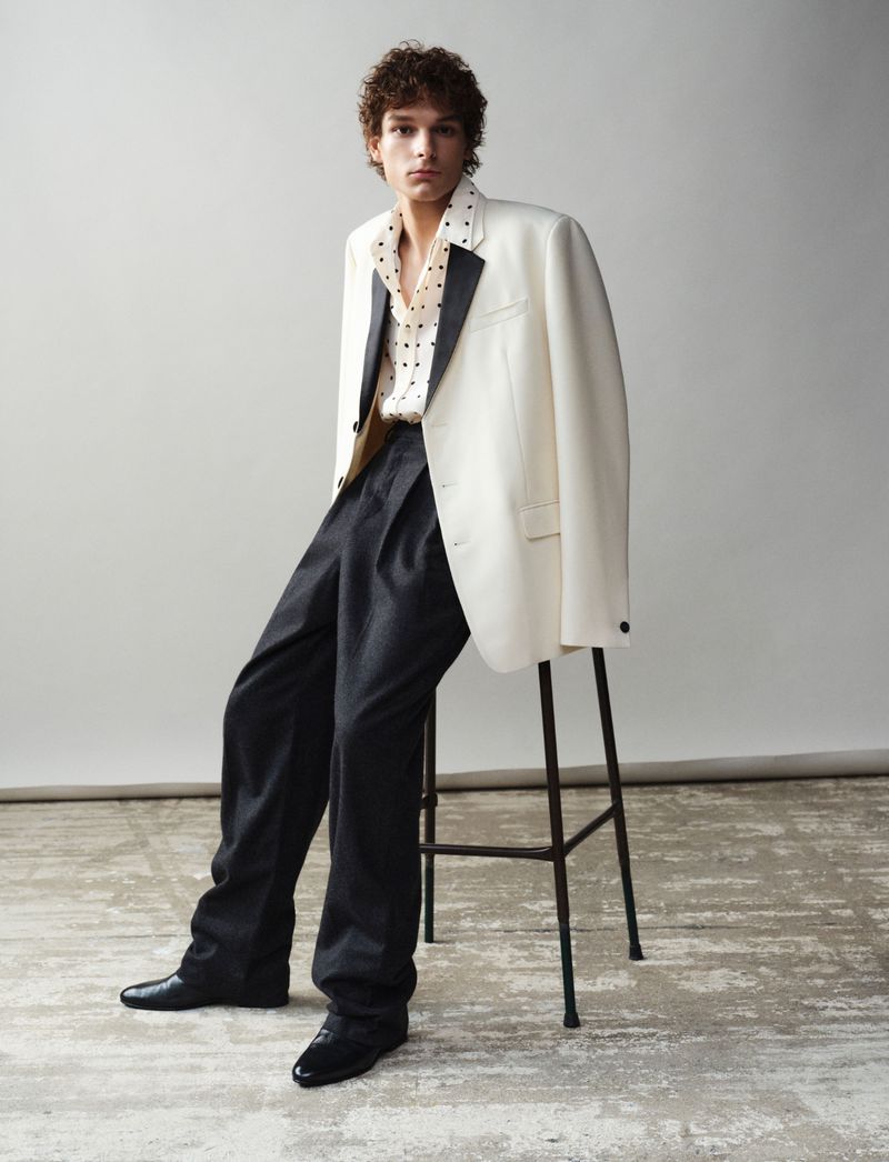 Man in ivory blazer and patterned shirt leaning on black stool in studio, looking at camera in fashion editorial-style shot.