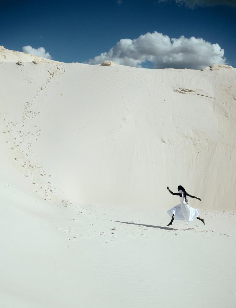 Woman in flowing white dress leaps on white sand dune beneath blue sky with scattered clouds in a fashion editorial style.