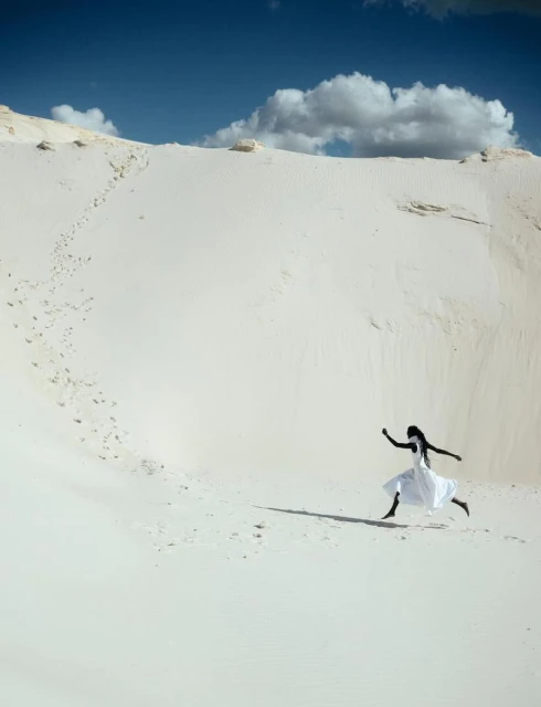 Woman in flowing white dress leaps on white sand dune beneath blue sky with scattered clouds in a fashion editorial style.