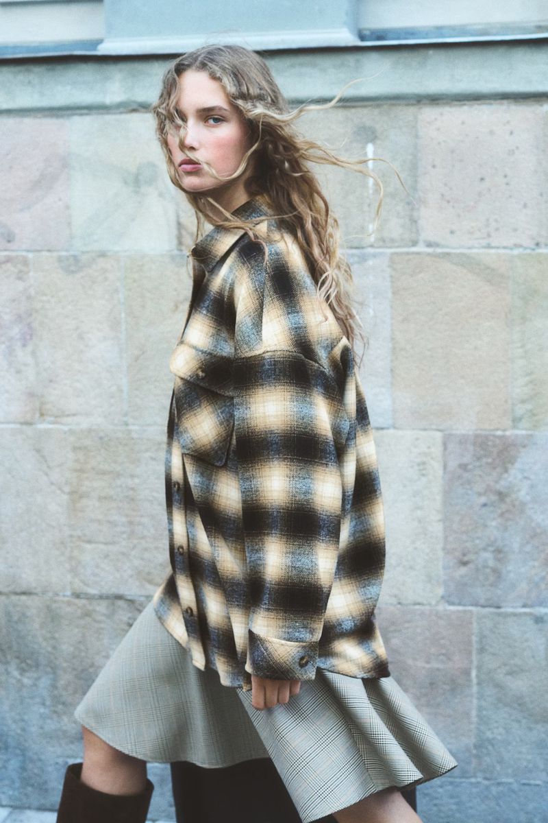 A young woman with blonde, wavy hair, wearing a plaid shirt jacket and skirt, walks past a stone wall on an overcast day.