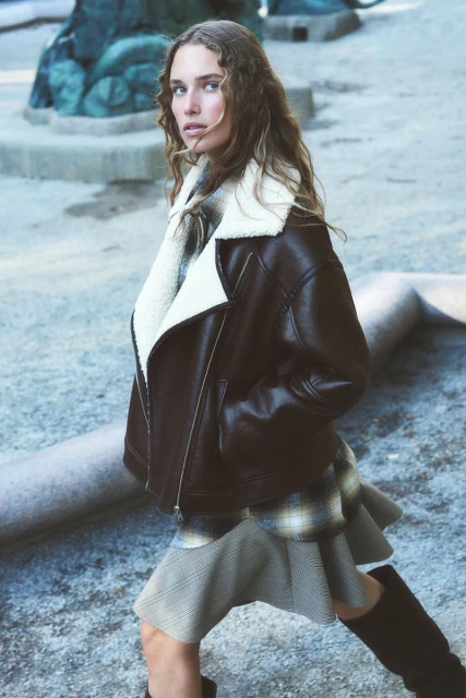 A woman with long brown hair models a brown leather shearling jacket and patterned skirt outdoors, posing near a stone sculpture.