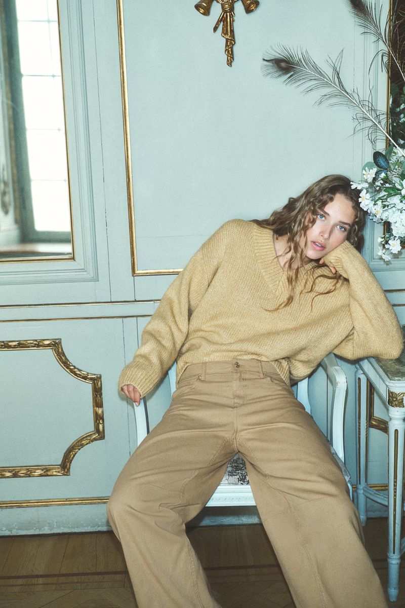 A woman with long curly hair poses in a tan sweater and wide-leg trousers, leaning on a chair against a light blue paneled wall.