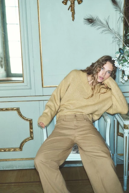 A woman with long curly hair poses in a tan sweater and wide-leg trousers, leaning on a chair against a light blue paneled wall.