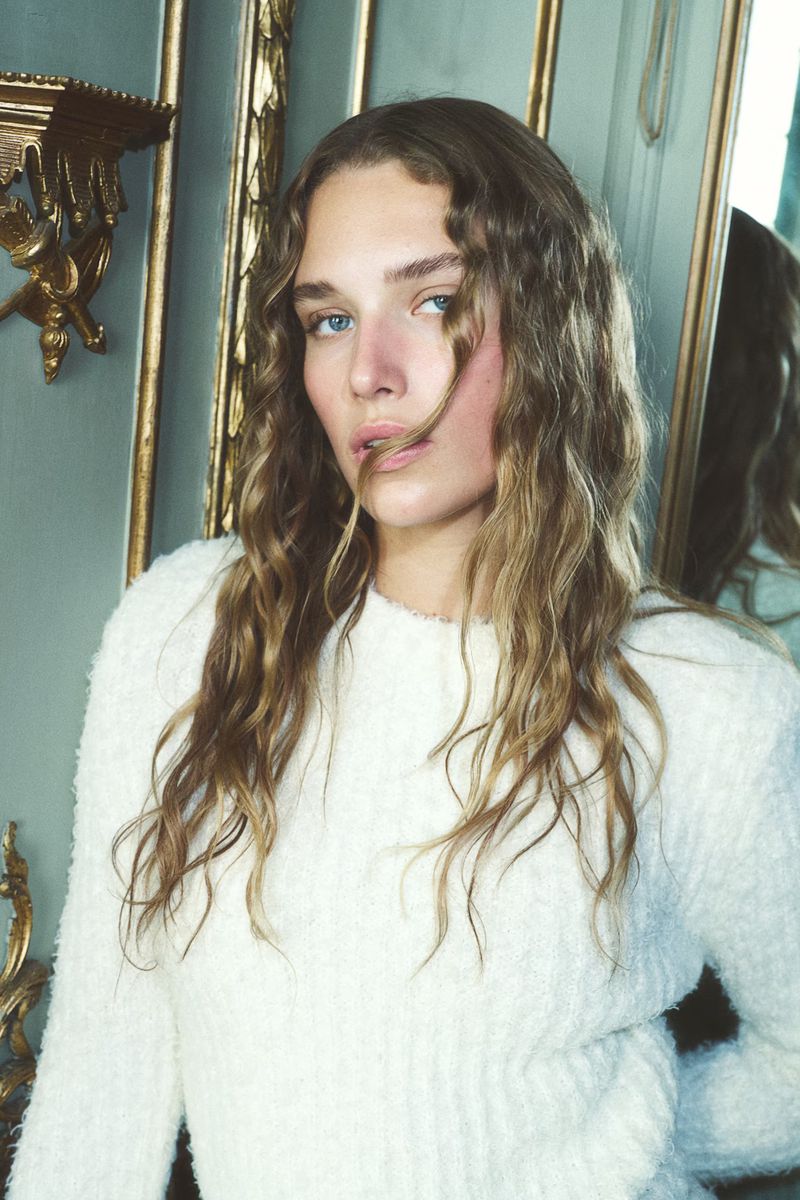 Portrait of a woman with wavy blonde hair wearing a white sweater, posing indoors with an ornate, neutral-colored background.