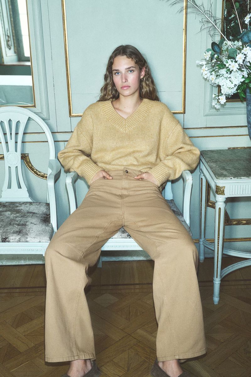 Indoor portrait of a female model in a light yellow sweater and wide brown pants, seated in a white chair, in a styled room with artwork.