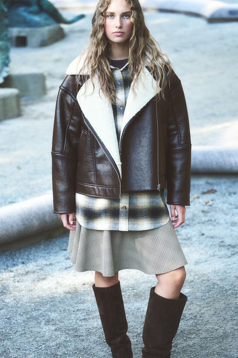 Fashion shot of a woman in a shearling-lined moto jacket, plaid shirt, skirt, and boots standing in an outdoor setting.