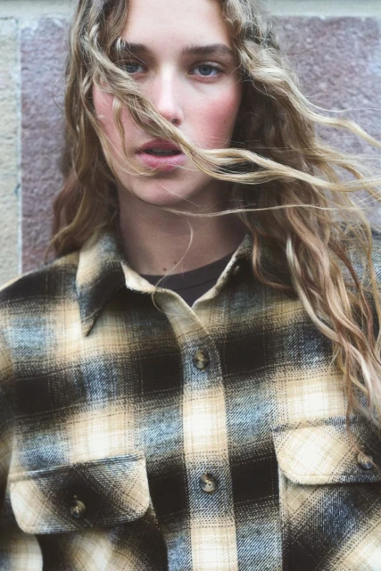 Close-up portrait of a young woman with wavy hair wearing a plaid shirt, posing against a textured wall. Fashion-focused image.