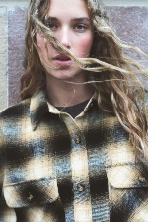 Close-up portrait of a young woman with wavy hair wearing a plaid shirt, posing against a textured wall. Fashion-focused image.