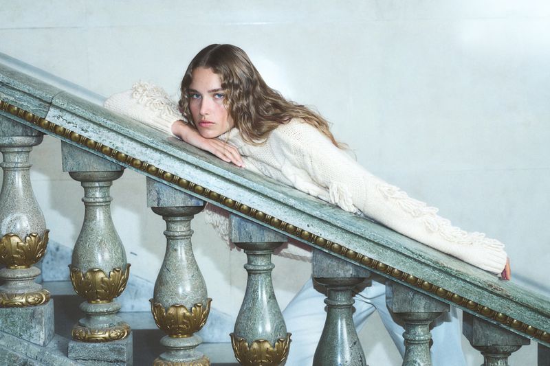 Woman in a cream-colored fringe sweater resting on a marble staircase, looking at the camera with a neutral expression. Indoor shot.