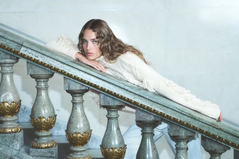 Woman in a cream-colored fringe sweater resting on a marble staircase, looking at the camera with a neutral expression. Indoor shot.