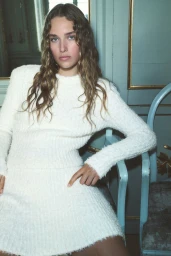 Model with long curly hair wearing a white textured sweater dress seated in a blue chair against a panelled wall