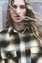 Close-up portrait of a female model with long blonde wavy hair wearing a plaid shirt against a stone wall