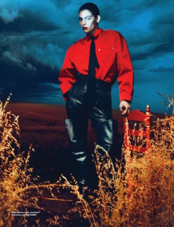 Person in red shirt and black pants posing in a field, with dramatic clouds and an antique chair in the outdoor setting.