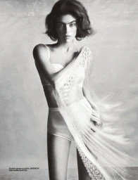 Model in white lingerie wearing a draped fringed wrap against a plain textured grey studio background