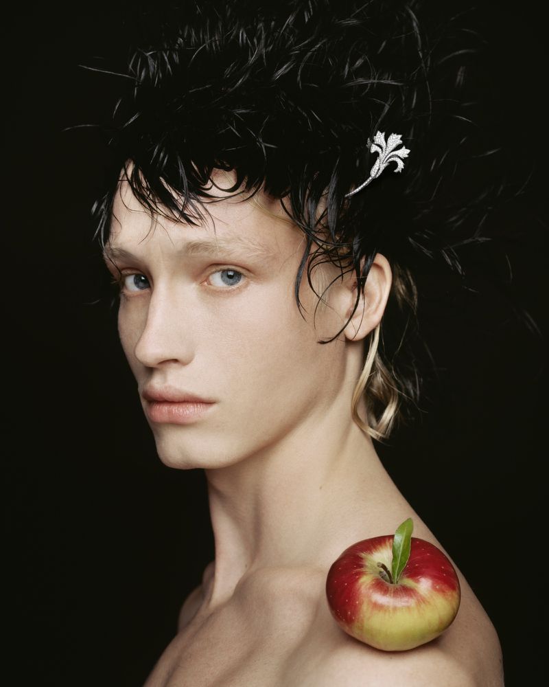 Young man with decorative headpiece and bare shoulders, posing in a studio with an apple resting on his shoulder against black backdrop.