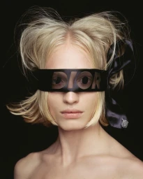 Model with blonde hair and a black Dior satin blindfold ribbon covering eyes against a black background.