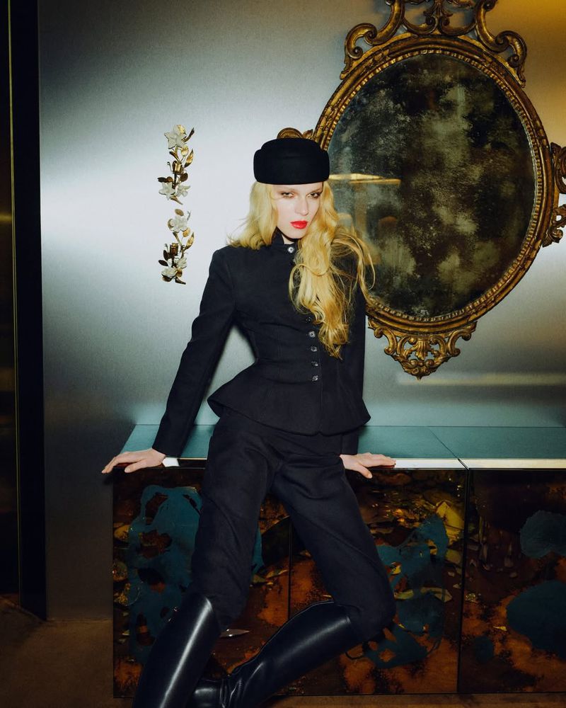 Elegant woman in black jacket, trousers, hat, and boots posing indoors with a vintage mirror and wall decor, beauty and fashion shot.