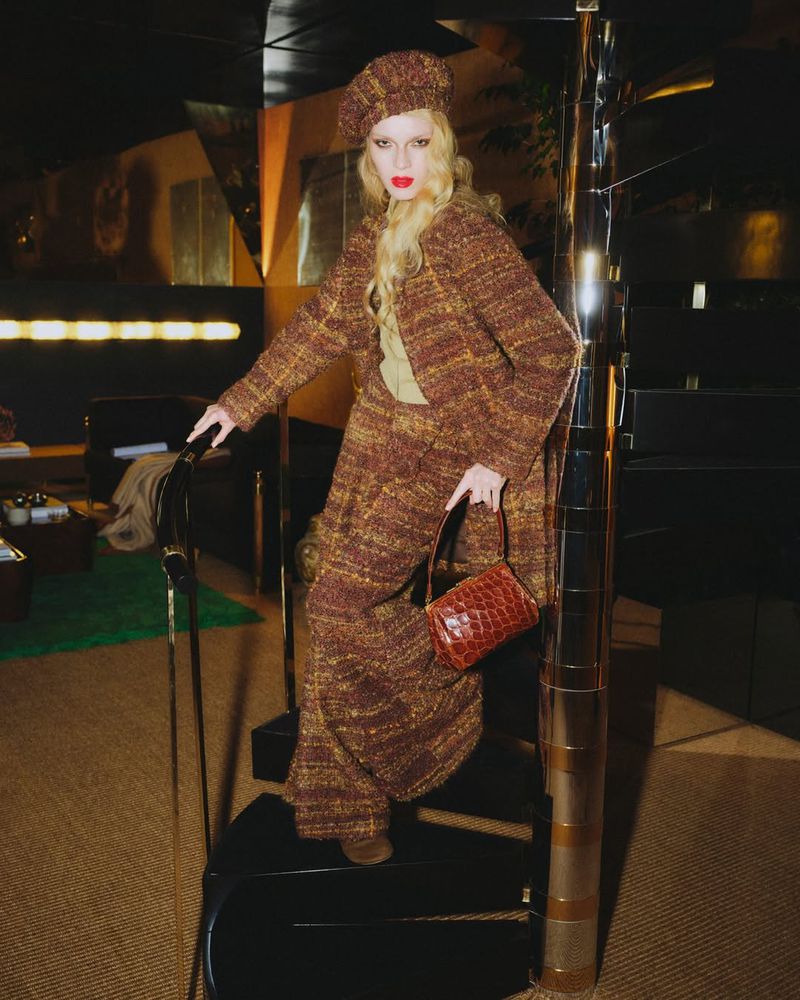 A long-haired fashion model in a matching brown tweed coat, beret, and wide-leg pants poses on a spiral staircase in a dark indoor environment.