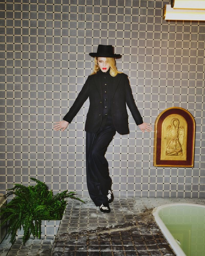Fashion shot of a young adult woman wearing a black blazer and hat, posing in a bathroom with tiled walls, a golden icon, and a tub.