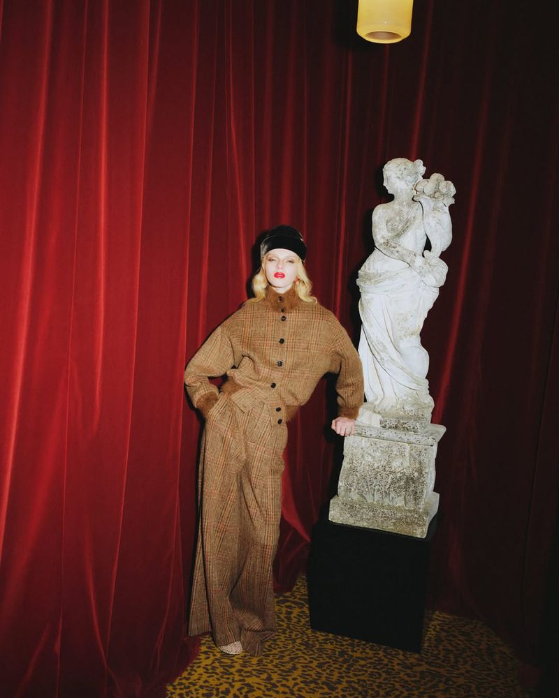 Fashionable woman in brown suit posing next to a classical statue in front of a red curtain in a studio setting, indoor lighting.