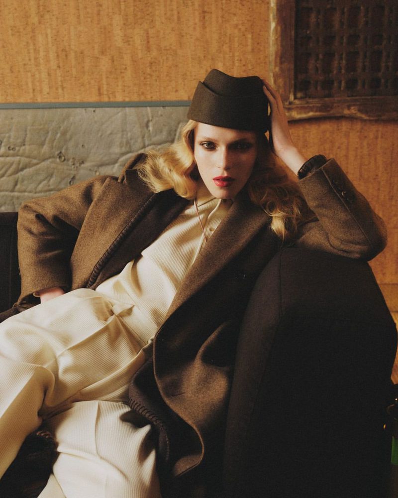 Woman in brown coat and hat sits with hand on her head indoors, featuring retro fashion and neutral colors, in a styled editorial shot.