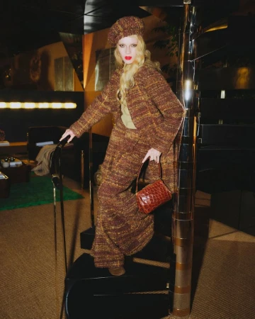 Model wearing a brown textured tweed suit and beret standing on a spiral staircase holding a crocodile-embossed handbag.