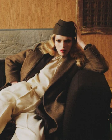 Model Colin Jones in a brown wool coat, cream trousers, and dark garrison cap reclining on a sofa for Vogue Italia.