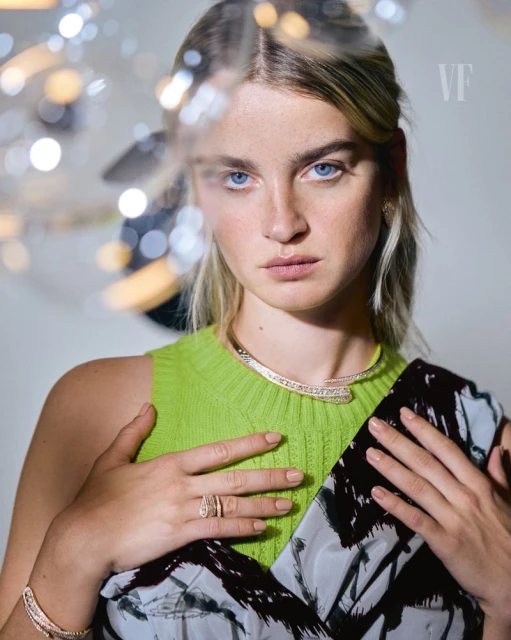 Blonde woman poses with jewelry, wearing a lime green sleeveless shirt with a printed dress overlay indoors, captured in a medium shot.